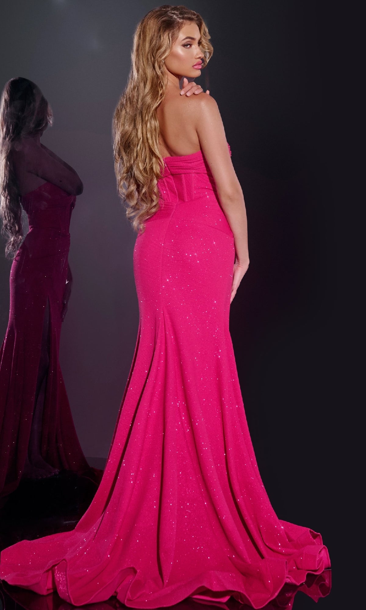 A woman with long wavy hair wears the Jovani 43065—a bright pink, floor-length, strapless glitter prom dress featuring a bow on the back—standing in front of a mirror reflecting her image.